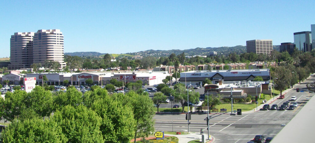 Aerial view of a shopping plaza with signboards, parking lots, and a busy intersection under a blue sky.
